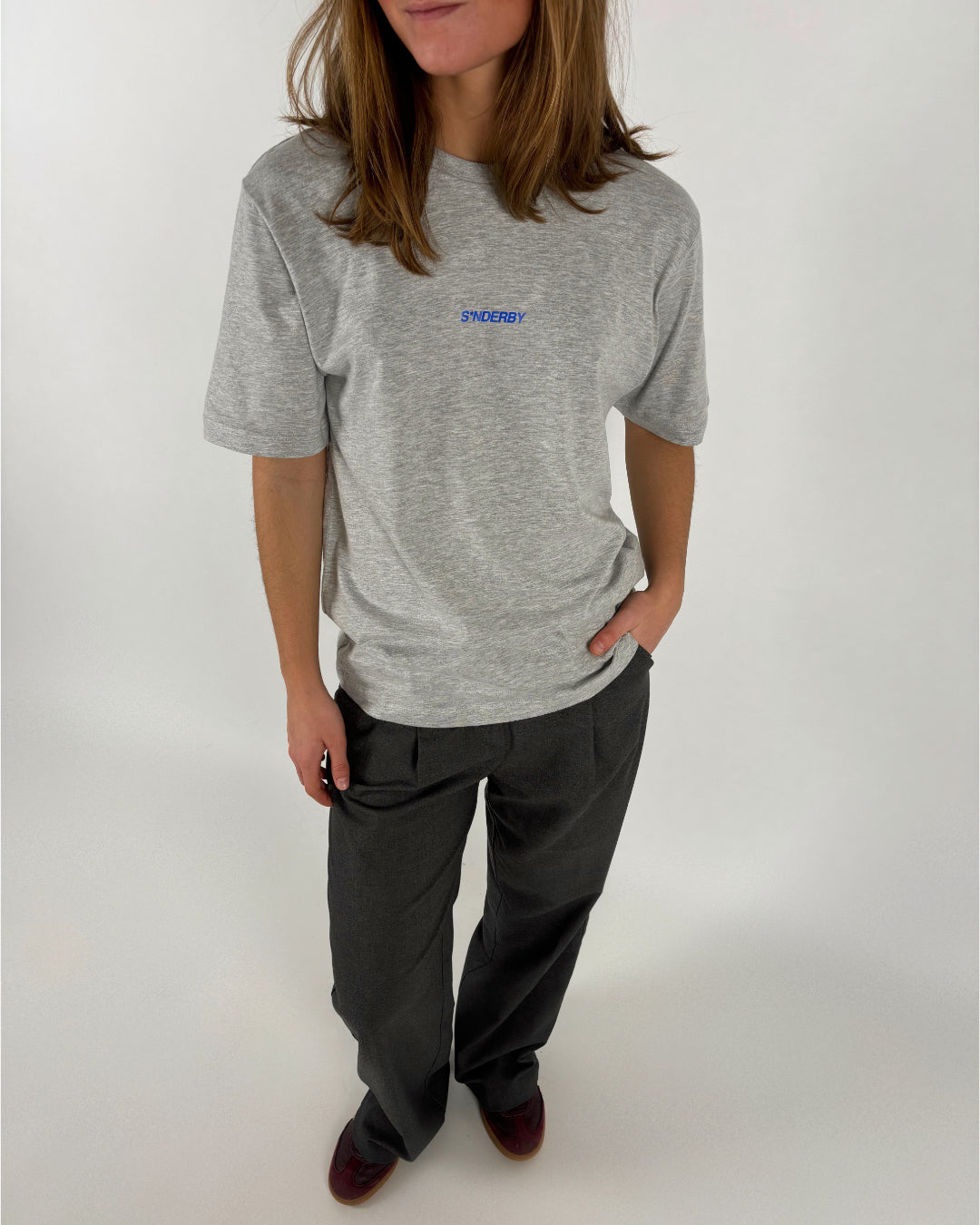 En person iført en grå T-shirt med ordet "SØNDERBY" på brystet står afslappet med hænderne i lommen, omgivet af en neutral, lys baggrund.