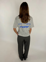 En person med langt hår bærer en grå T-shirt med blå tekst "SØNDERBY 301103" og yderligere tekst i et studie med hvid baggrund.