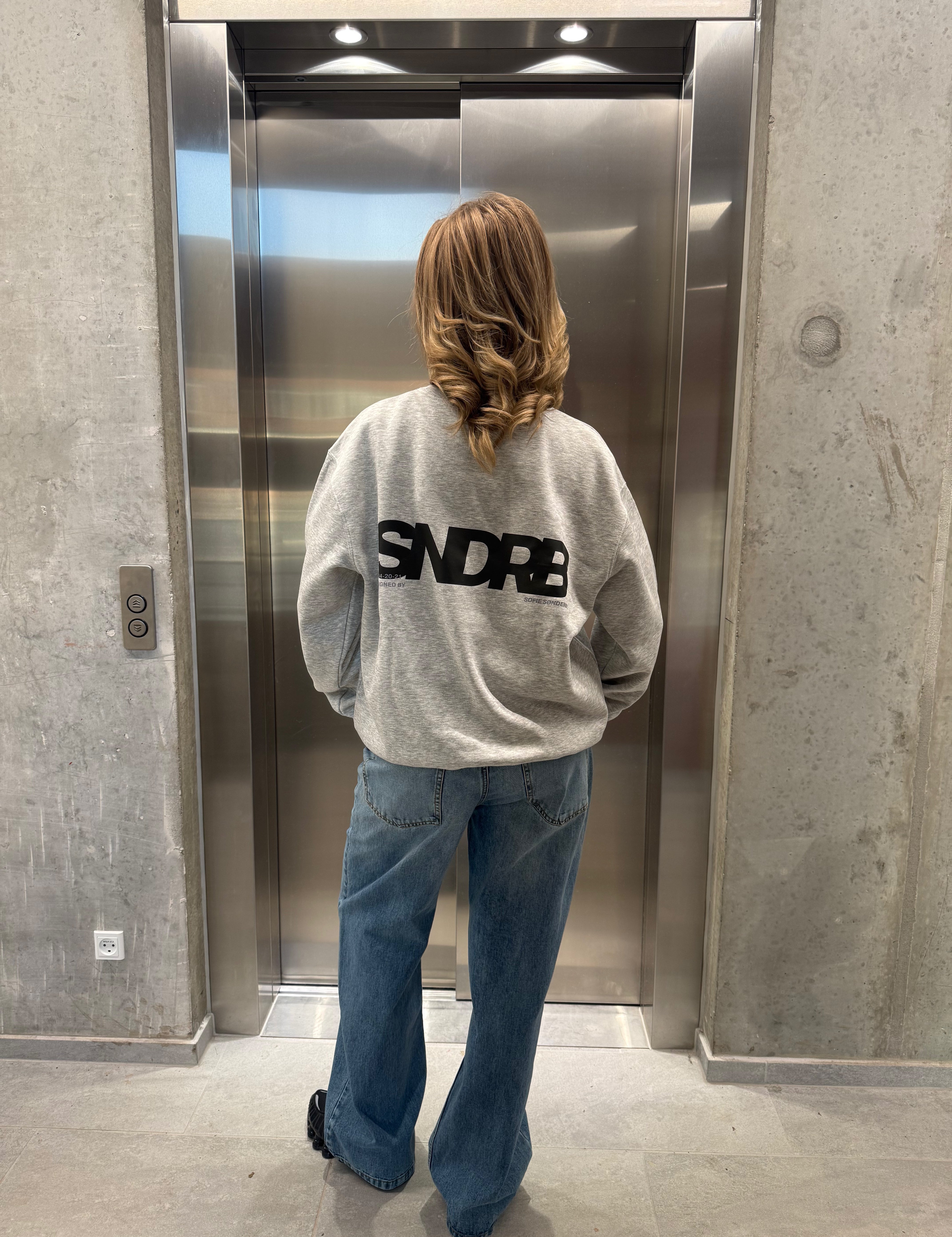 Stemningsbillede af pige der bærer en grå sweatshirt med sort SNDRB tryk og blå NANNA jeans.  Fra BySofieSønderby