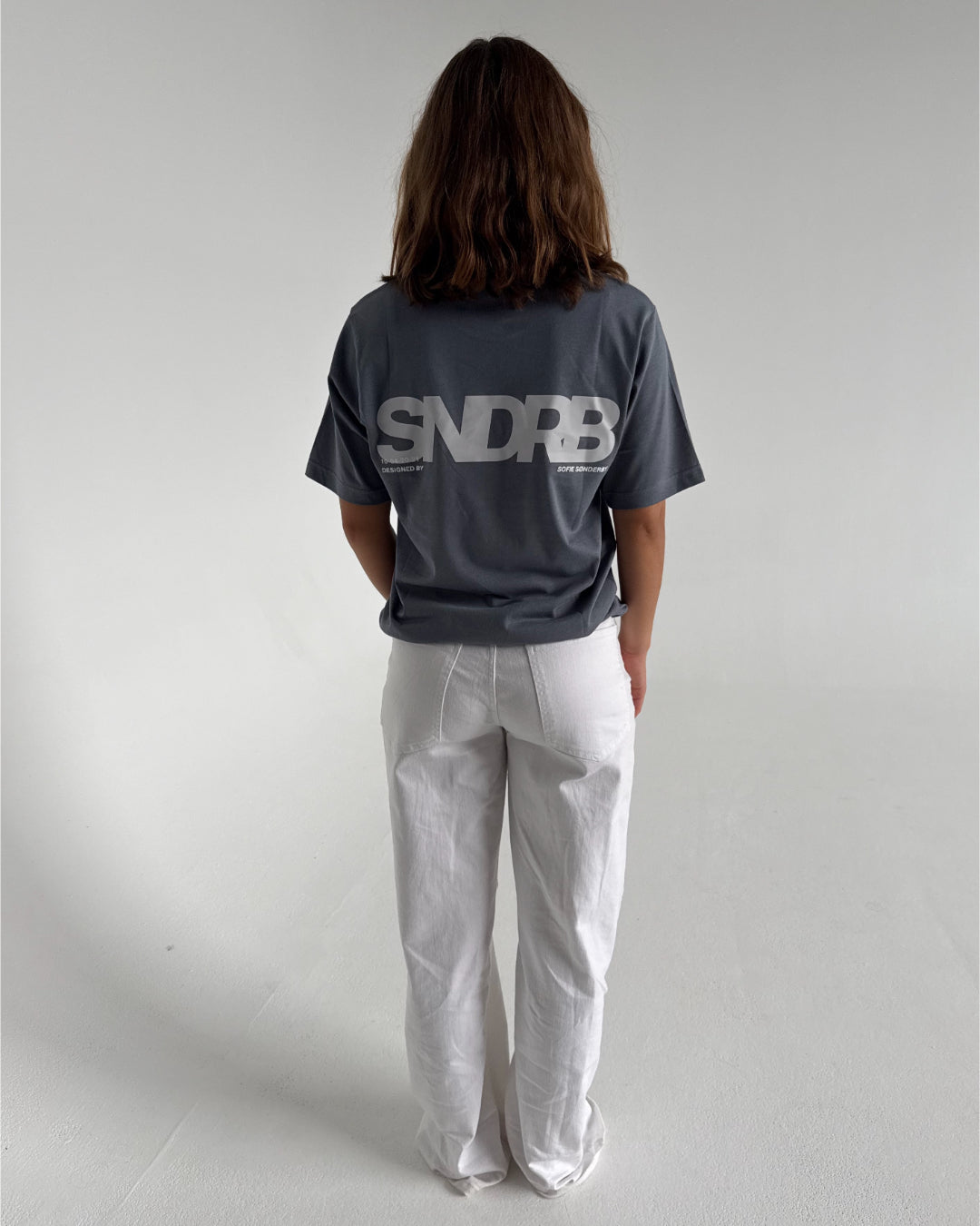 Modelfoto af pige som bærer en SNDRB tee i farven stone, samt et par hvide jeans. Alt fra By Sofie Sønderby