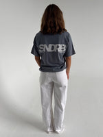 Modelfoto af pige som bærer en SNDRB tee i farven stone, samt et par hvide jeans. Alt fra By Sofie Sønderby