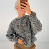 En person tager en spejlslefie med en mobiltelefon, iført en grå, strikket sweater og brune bukser. Baggrunden er simpel og lys.