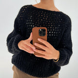 Person tager et spejlbillede med en smartphone. Personen bærer en sort strikket sweater og en brun bukser, med et enkelt guldsmykke om håndleddet. Baggrunden er enkel og lys.