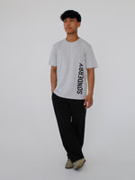 Person står iført en grå t-shirt med teksten "SØNDERBY", sorte bukser og beige sko mod en neutral baggrund.