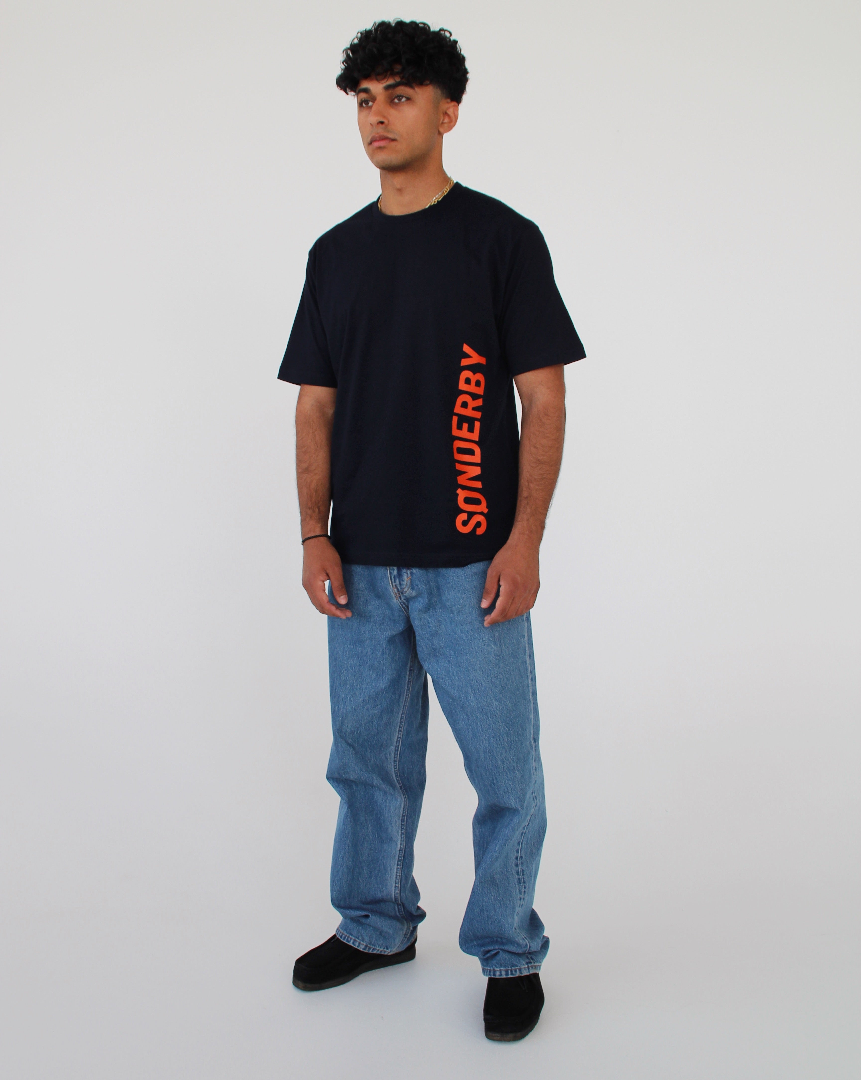 En mand står i afslappet tøj; han bærer en sort t-shirt med ordet "SØNDERBY" skrevet lodret i orange. Han er i et minimalistisk miljø uden distraktioner.