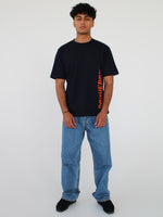 En person står i et hvidt rum, iført en sort T-shirt med orange tekst "CONQUER" og lyseblå jeans. Personen ser fremad med armene afslappet ned langs siderne.