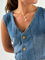 En person iført en denim vest med knapper, bærer to delikate halskæder med små ovale vedhæng. Personen står mod en neutral baggrund.