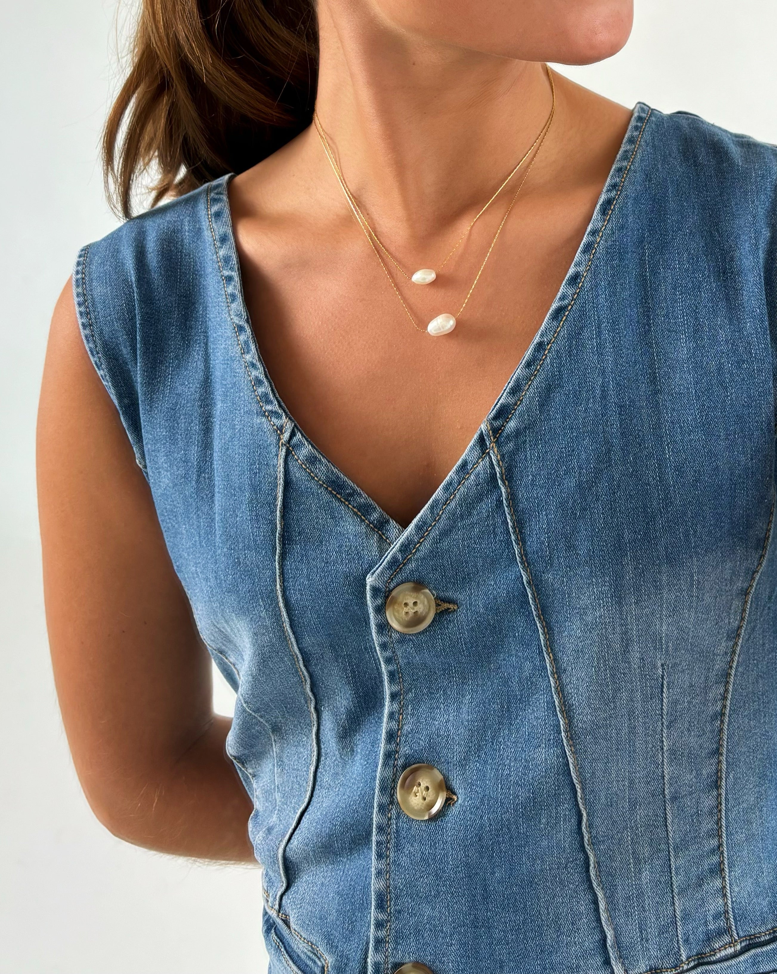 En person iført en denim vest med knapper, bærer to delikate halskæder med små ovale vedhæng. Personen står mod en neutral baggrund.