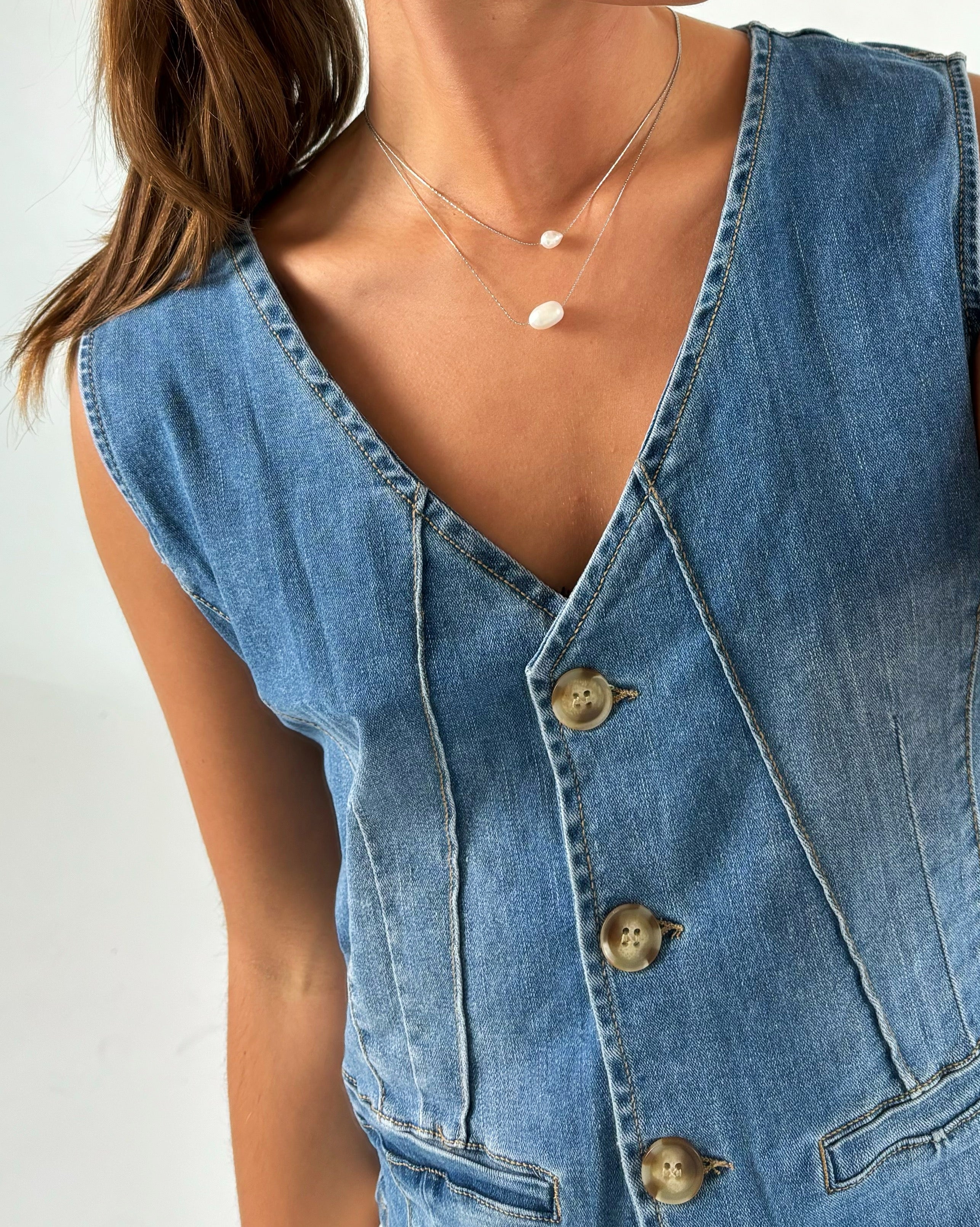 Person iført en blå denimvest med knapper, bærer en halskæde med to perler. Konteksten er en lys baggrund, og vesten har synlige sømme og lommer.
