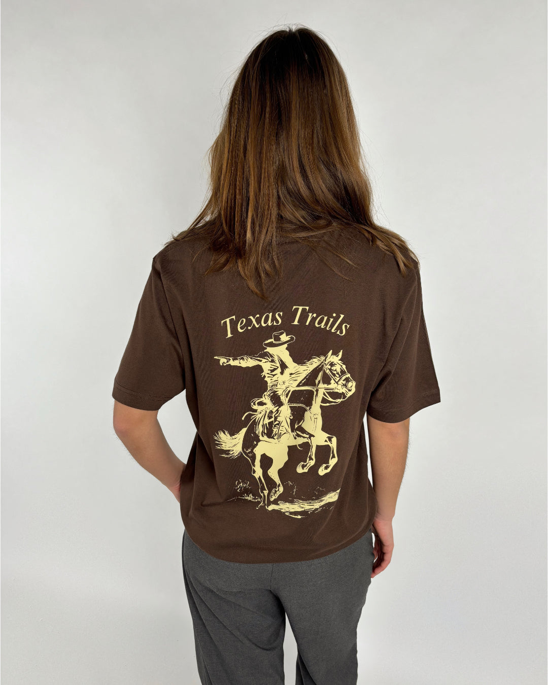 En person iført en brun T-shirt med påtrykt motiv af en cowboy på en hest med teksten "Texas Trails". Personen står mod en neutral, lys baggrund.