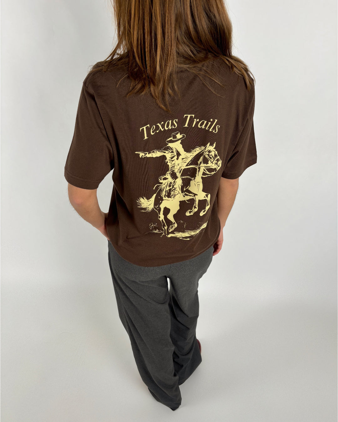 En person med langt hår bærer en brun t-shirt med teksten "Texas Trails" og en illustration af en cowboy på hesteryg i en lys nuance står i et neutralt rum.
