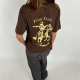 En person med langt hår bærer en brun t-shirt med teksten "Texas Trails" og en illustration af en cowboy på hesteryg i en lys nuance står i et neutralt rum.