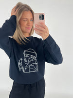 En person tager et selfie med en lyserød telefon og blinker. De bærer en sort trøje med en stregtegning af en person, der holder en flaske.