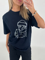 Kvinde i sort t-shirt med line art-tryk af person, holder mobiltelefon op til spejl. Hun står i minimalistisk, hvidt rum iført perlearmbånd.