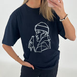Kvinde iført sort T-shirt med en hvidt lineart design af en person med hætte og flaske. Hun ser sig i spejlet med en mobiltelefon i et lyst rum.