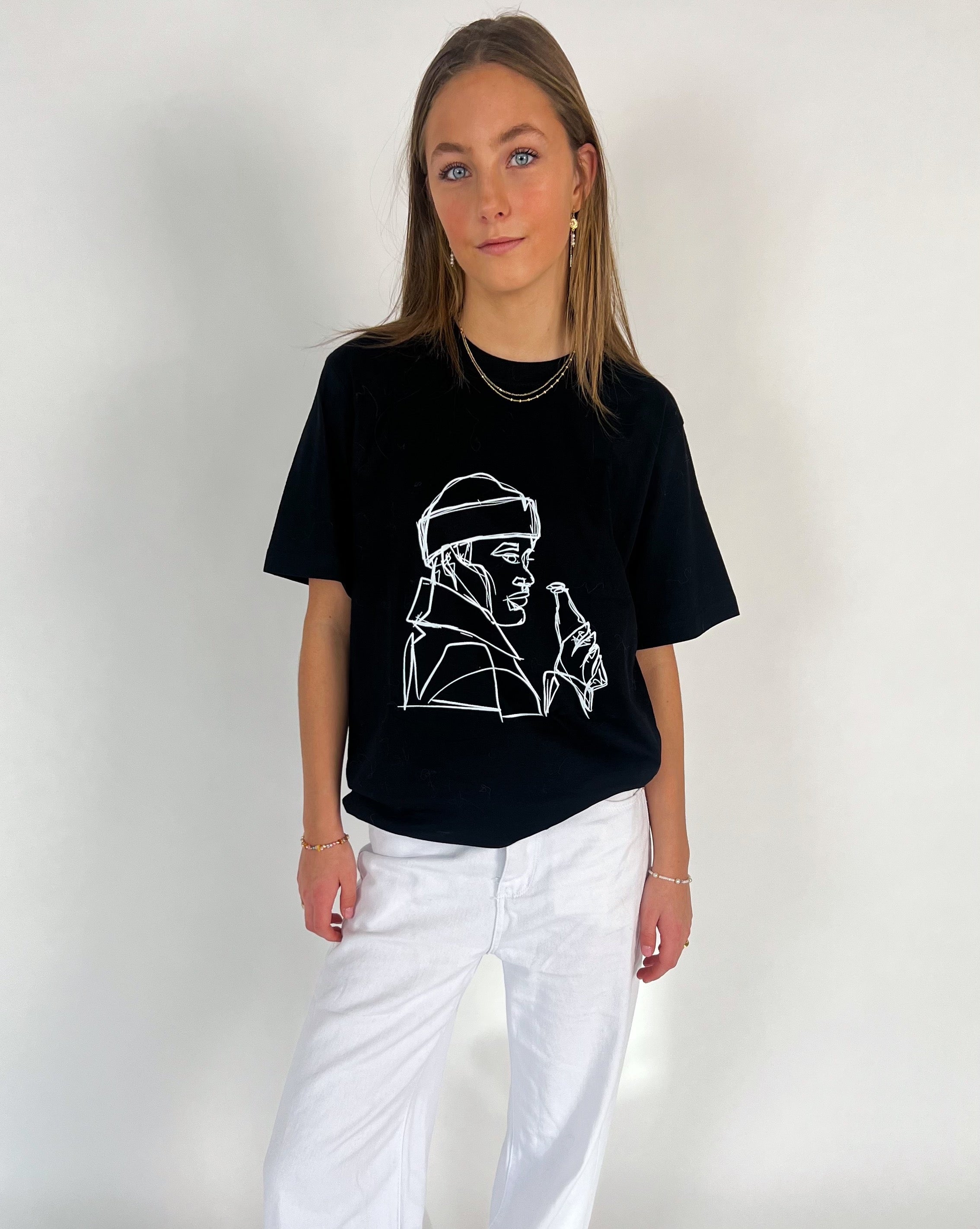 En person iført en sort t-shirt med hvidt lineart-motiv og hvide bukser står i et lyst rum. Vedkommende har langt hår og bærer smykker.