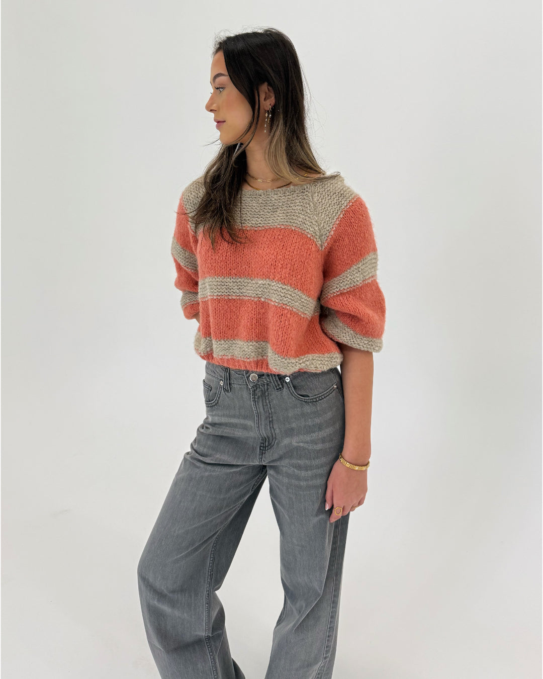 En kvinde står og kigger til siden. Hun bærer en stribet trøje i orange og beige samt grå jeans. Baggrunden er hvid og minimalistisk.
