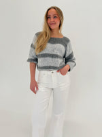 En person iført en gråstribet sweater og hvide bukser står afslappet med en hånd i lommen mod en lys baggrund.