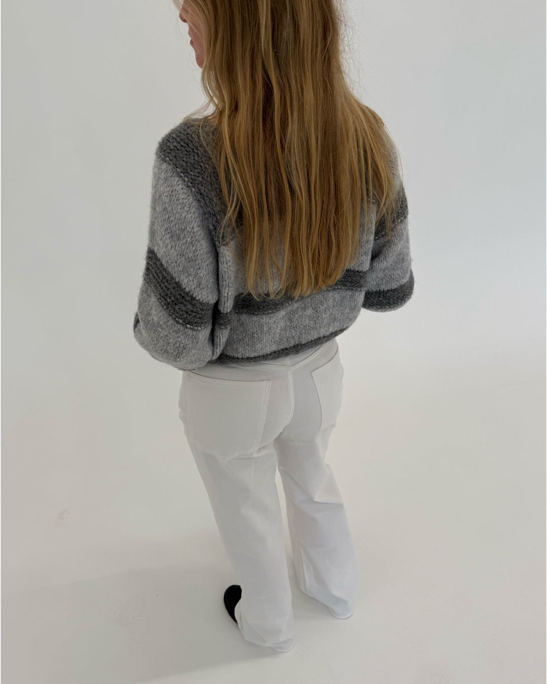 En person med langt hår står med ryggen til, iført en gråstribet sweater og hvide bukser, i et minimalistisk, lyst rum.