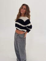 Kvinde i en sort-hvid stribet sweater og grå jeans står afslappet med hånden i lommen, smilende i et lyst rum med en hvid baggrund.