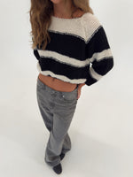 En person iført en sort-hvid stribet sweater står med hænderne i lommerne på et par grå bukser i et lyst studie.