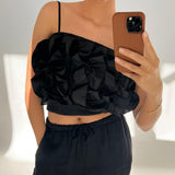 En person iført sort rynket top og bukser tager en selfie med en brun mobiltelefon i et lyst rum. Personen bærer et armbånd og står med ene hånd i lommen.
