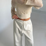 En person iført en hvid, åbenstrikket sweater samt hvide bukser står i et minimalistisk rum med lyse vægge og en hvid gulvflade.