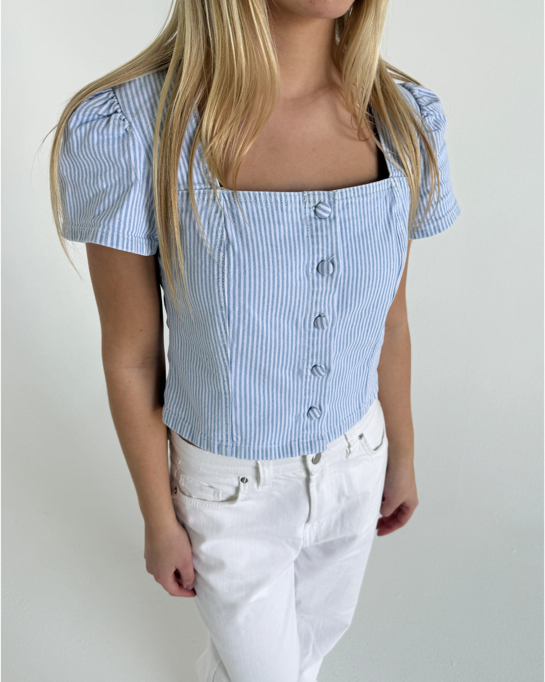 Blond kvinde iført en blå- og hvidstribet skjorte med knapper og hvidt jeans, står mod neutralt, hvidt baggrund.