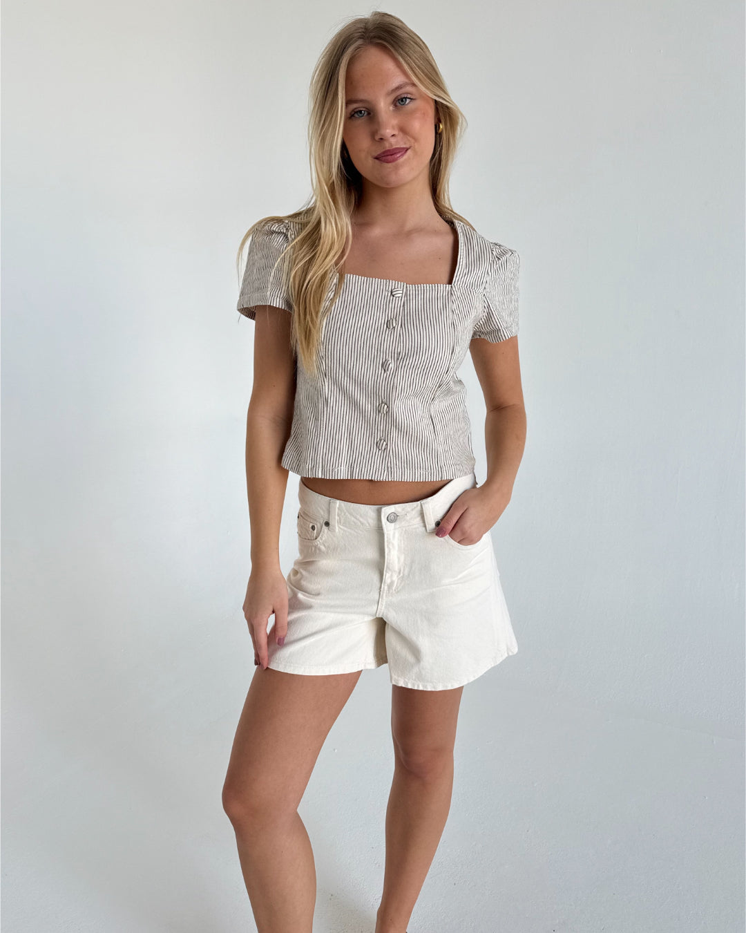 En kvinde står afslappet i en hvidblå stribet top og hvide shorts i et minimalistisk rum med lyse vægge.