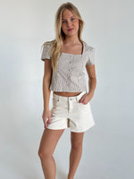 En kvinde står afslappet i en hvidblå stribet top og hvide shorts i et minimalistisk rum med lyse vægge.