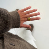 Hånd bærer en gylden ring og peger nedad. Personen har en brunt sweater og hvide bukser på. Støvler er synlige i baggrunden på en hvid baggrund.