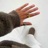 En hånd med en brun sweater bærer to store ringe, én guld og én sølv. Baggrunden viser hvide bukser og en brun sko på et lyst gulv.