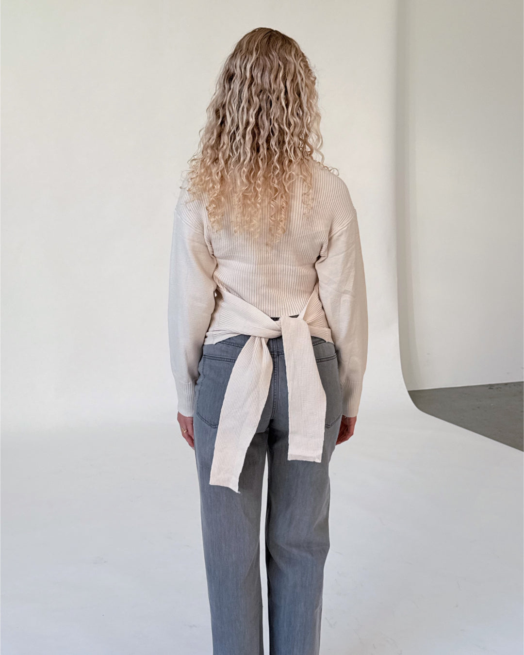 Modelfoto af pige i beige WHITNEY strik, samt grå NANNA jeans. Alt er fra BySofieSønderby.