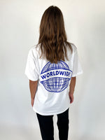 En person iført en hvid t-shirt med et blåt globusmotiv og teksten "WORLDWIDE" står med ryggen til kameraet i et neutralt, lyst rum.